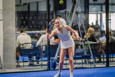 Riga, Letonya - 3 Mart 2024 - Beyaz elbiseli bir bayan padel oyuncusu, kollarını sarı bir topa raketiyle vurmak için uzatıyor, kapalı alanda, arka planda seyirciler.