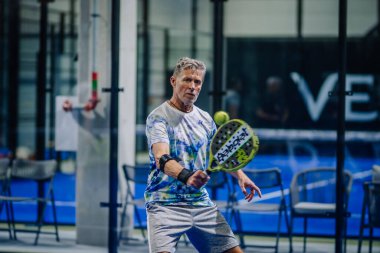 Riga, Letonya - 3 Mart 2024 - Gri saçlı olgun bir erkek padel oyuncusu yeşil bir raketle forehand vurmak üzere, kravatlı, odaklanmış.