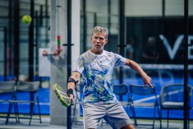 Riga, Letonya - 3 Mart 2024 - Renkli gömlek, şort ve bileklik giymiş, forehand vuruşuna hazırlanan bir adam Padel oynuyor..