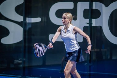 Riga, Letonya - 3 Mart 2024 - Beyaz giysili odaklı bir bayan padel oyuncusu büyük harflerle arkaplanda siyah beyaz raketle topa vurmaya hazır.