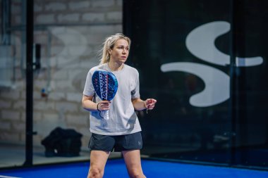 Riga, Letonya - 3 Mart 2024 - Bir kadın padel kortunda elinde mavi bir kürekle dikiliyor, sıradan bir tişört ve şort giyiyor, odaklanmış bir bakışla, camdan görünen büyük harflerle..