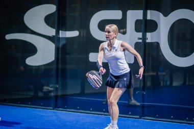 Riga, Letonya - 3 Mart 2024 - Siyah etekli beyaz spor elbiseli bir kadın arka planda büyük beyaz harflerle odaklanmış bir ön çekim için Padel çalıyor..