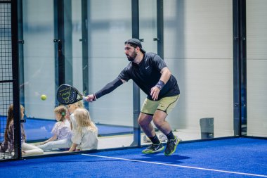 Riga, Letonya - 3 Mart 2024 - Spor giyimli sakallı bir adam kapalı mavi bir sahada raketiyle padel topuna vurmaya hazırlanıyor..