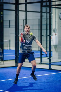 Riga, Letonya - 3 Mart 2024 - Gri spor gömlekli ve şortlu odaklanmış bir adam padel oynuyor, kapalı bir mavi kortta topa vurmaya hazır.
