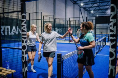 Riga, Letonya - 3 Mart 2024 - Padel kortunda üç kadın, beyaz giyen iki kadın yürürken, yeşil ve siyah giyen üçüncüsü beşlik çakıyor ve sportmenlik sergileniyor.