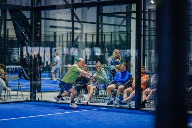 Riga, Letonya - 3 Mart 2024 - Sarı tişörtlü bir adam, mavi bir kortta, cam bir çitin arkasında oturup maçı yakından izleyen seyircilerle bir çekim için Padel oynar..