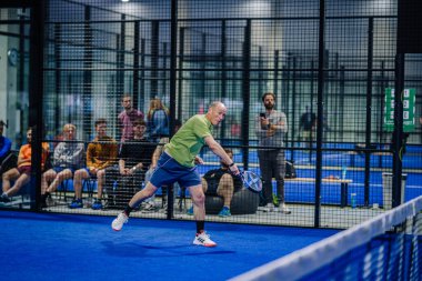 Riga, Letonya - 3 Mart 2024 - Yeşil bluzlu ve donanma şortlu bir oyuncu mavi raketle düşük padel atış yapmak için gerinirken, seyirciler mahkemenin çitinin arkasından izliyor