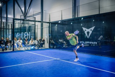 Riga, Letonya - 3 Mart 2024 - Sarı tişörtlü bir adam padel oynuyor, mavi bir korta ters vuruş yapıyor, seyirci cam bir duvarın arkasında..