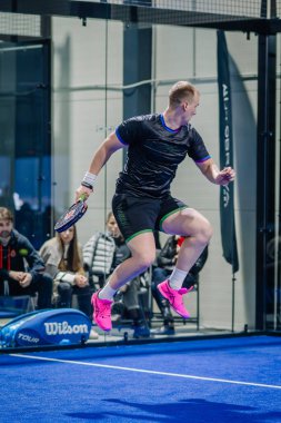 Riga, Letonya - 3 Mart 2024 - Siyah spor giyim ve parlak pembe ayakkabılı bir oyuncu padel topuna vurmak için sıçrıyor..