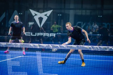 Riga, Letonya - 3 Mart 2024 - İki oyuncu mavi bir padel kortunda; biri önde voleybol için hazır, diğeri arkada, 'CUPRA' markası görünür