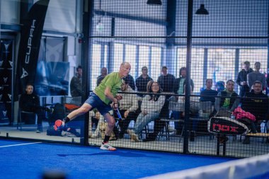 Riga, Letonya - 3 Mart 2024 - Seyirciler yoğun maçı çitten izlerken bir oyuncu sahanın üzerinde düşük bir padel topuna ulaşmak için esniyor..