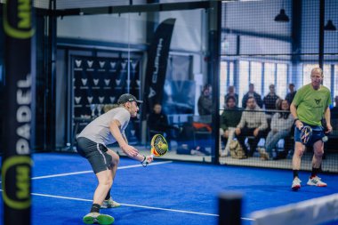 Riga, Letonya - 3 Mart 2024 - İki adam Padel oynuyor; biri ön planda topa vurmaya hazır, diğeri arka planda mavi bir kortta, seyirciler izliyor