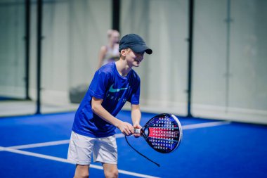 Riga, Letonya - 3 Mart 2024 - Mavi tişörtlü ve beyaz şortlu genç bir oyuncu, elinde raketle arka planda başka bir oyuncuyla Padel kortunda gülümsüyor ve oynamaya hazır..