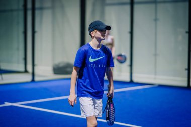 Riga, Letonya - 3 Mart 2024 - Mavi Nike tişörtlü ve şapkalı bir genç, padel raket tutuyor, bir turnuva sırasında Padel kortunda odaklanmış bir ifadeyle.