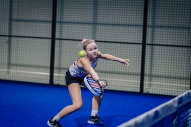 Riga, Letonya - 3 Mart 2024 - Genç bir padel oyuncusu, yaklaşan topa odaklanmış, ters vuruş için hazır, kolsuz bluz giyiyor ve mavi bir kortta şort giyiyor