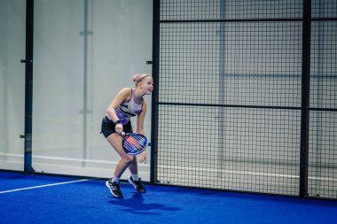 Riga, Letonya - 3 Mart 2024 - Odaklanmış bir oyuncu bir padel maçında düşük bir topa vurmaya hazırlanıyor, mavi bir kortta desenli bir atlet ve siyah şort giyiyor.