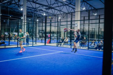Riga, Letonya - 3 Mart 2024 - İç sahada iki adam Padel oynuyor, biri yukarıdan şut atıyor. Seyirciler, geniş bir salonda, parlak ışıkların altında, cam bir camın arkasından izlerler..