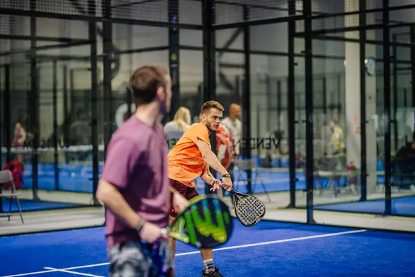Riga, Letonya - 3 Mart 2024 - Kapalı bir tesiste iki adam padel oynuyor; turuncu gömlekli biri raketiyle topa vurmaya hazır.