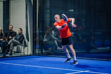 Riga, Letonya - 3 Mart 2024 - Turuncu tişörtlü bir adam arka planda seyirci ile arka planda vuruşunu yapmak üzere Padel oynuyor.