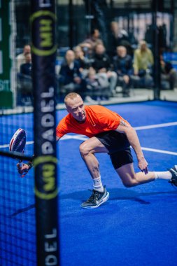 Riga, Letonya - 3 Mart 2024 - Turuncu tişörtlü bir erkek padel oyuncusu, arka planda seyirciler olan mavi bir kortta kürekle topa vurmak için geriniyor.