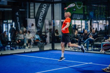 Riga, Letonya - 3 Mart 2024 - Turuncu bluzlu bir oyuncu, arka planda seyircilerle birlikte bir Padel maçında backhand vuruşu yaparken havada uçuyor.