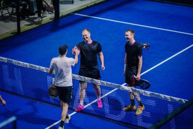 Riga, Letonya - 3 Mart 2024 - Padel kortunda üç erkek ağda; beyaz gömlekli biri maç veya puandan sonra diğerlerini selamlıyor veya tebrik ediyor gibi görünüyor.