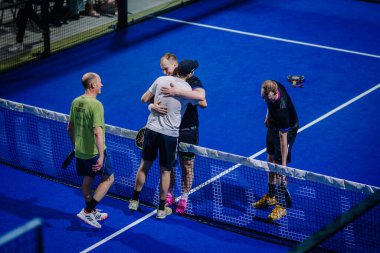 Riga, Letonya - 3 Mart 2024 - Padel kortunda oynayan dört oyuncu maç sonrası etkileşime girer, iki oyuncu ağda sarılır ve diğer ikisi de.