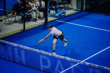 Riga, Letonya - 3 Mart 2024 - Eylemin ortasında bir adam arka planda seyircilerin olduğu kapalı mavi bir kortta raketle ağlara doğru atlıyor..