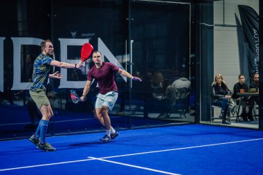 Riga, Letonya - 3 Mart 2024 - İki adam padel oynuyor; biri ön planda kırmızı raketle topa vurmaya hazır, seyirciler cam duvarların arkasından izliyor