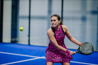 Riga, Letonya - 3 Mart 2024 - Pembe leopar desenli elbiseli bir kadın padel çalıyor, havada bir topa odaklanmış, elinde siyah bir padel raketi, kapalı mavi bir kortta.