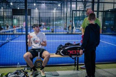 Riga, Letonya - 3 Mart 2024 - Padel Court 'ta elinde bir şişe ve jöle ile bir bankta oturan bir adam kameraya arkası dönük duran biriyle konuşuyor..