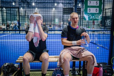 Riga, Letonya - 3 Mart 2024 - Padel kortunda iki adam oturuyor; biri yüzünü havluyla kurutuyor, diğeri su şişesi tutuyor.