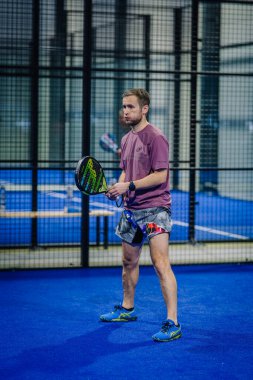 Riga, Letonya - 3 Mart 2024 - Padel raket tutan bir adam, mavi padel bir kortta leylak rengi bir gömlek, baskılı şort ve mavi spor ayakkabılar giyiyor. Arka planda bir kafes görünüyor..