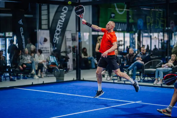 Riga, Letonya - 3 Mart 2024 - Turuncu bluzlu bir oyuncu, arka planda seyircilerle birlikte bir Padel maçında backhand vuruşu yaparken havada uçuyor.