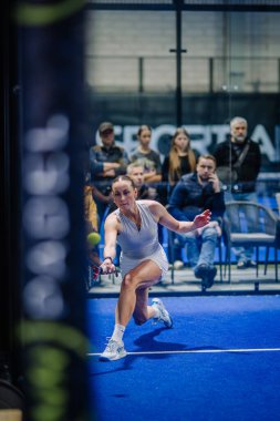Riga, Letonya - 3 Mart 2024 - Beyaz elbiseli bir kadın padel oyuncusu mavi bir salonda düşük bir atış yapmak için harekete geçti, arka planda seyirciler bulanıklaştı.