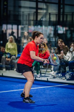 Riga, Letonya - 3 Mart 2024 - Kırmızı bluzlu bayan bir padel oyuncusu topa odaklanmış, vurmaya hazır, arka planda mavi sahada bir kalabalık var.