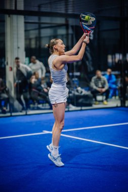 Riga, Letonya - 3 Mart 2024 - Beyaz kıyafetli odaklanmış bir bayan padel oyuncusu maç sırasında arka planda seyircilerin olduğu yüksek bir atış için uzanıyor..