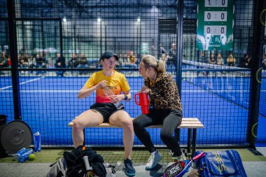 Riga, Letonya - 3 Mart 2024 - Mahkemenin yanındaki bankta dinlenen ve etkileşime giren iki padel oyuncusu, arkalarında teçhizat ve seyirci ile birlikte.
