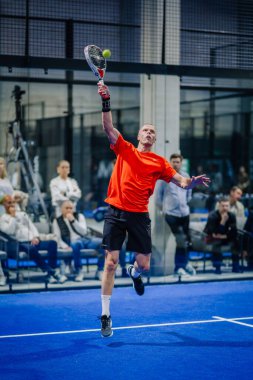 Riga, Letonya - 3 Mart 2024 - Turuncu tişörtlü bir erkek oyuncu bir padel maçında yüksek topa vurmak için sıçrıyor..