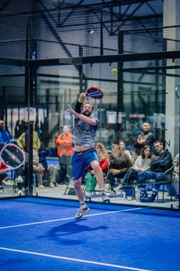 Riga, Letonya - 3 Mart 2024 - Spor giyim sektöründe sakallı bir adam arka planda seyirci ile bir padel maçında topa vurmak için zıplar.