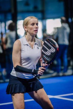 Riga, Letonya - 3 Mart 2024 - Beyaz kolsuz bluzlu ve siyah etekli bir kadın Padel raket tutuyor, sahanın dışına odaklanmış bir ifadeyle bakıyor.