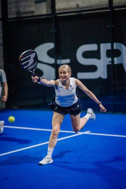 Riga, Letonya - 3 Mart 2024 - Beyaz atletli ve siyah etekli bir kadın padel oynuyor, kollarını kapalı bir mavi kortta topa uzanmak için uzatıyor.