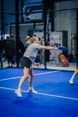 Riga, Letonya - 3 Mart 2024 - Kadın bir padel oyuncusu arka planda seyircilerin olduğu kapalı mavi bir sahada ters atış yapmak için kolunu uzatır.