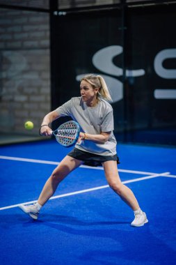 Riga, Letonya - 3 Mart 2024 - Kadın bir Padel oyuncusu, yaklaşan topa odaklanmış, iç saha mavi kortunda ters vuruş yapmaya hazır.