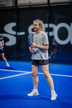 Riga, Letonya - 3 Mart 2024 - Gülümseyen bir bayan padel oyuncusu arka planda başka bir oyuncuyla mavi bir kortta raket tutuyor.