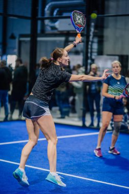 Riga, Letonya - 3 Mart 2024 - Odaklanmış bir bayan padel oyuncusu, arka planda başka bir bayan oyuncuyla birlikte kapalı mavi bir kortta forehand vurmak üzere..