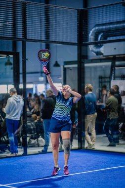 Riga, Letonya - 3 Mart 2024 - Bir bayan padel oyuncusu bir maçta topa vurmak için yükseğe uzanır, arka planda seyirciler, kapalı mavi bir kortta.