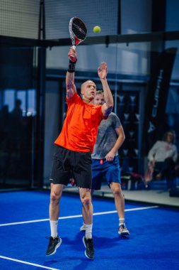 Riga, Letonya - 3 Mart 2024 - Turuncu tişörtlü odaklanmış bir oyuncu, mavi kortta oynanan çiftler maçında topa vurmak için uzatır..