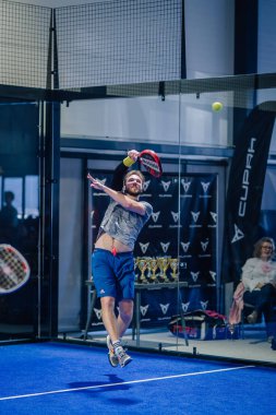 Riga, Letonya - 3 Mart 2024 - Sakallı bir adam padel oynuyor, yüksek topa vurmak için zıplıyor, arka planda kupalar ve markalar var.