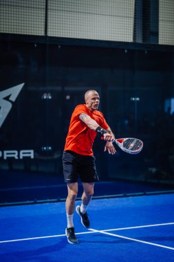 Riga, Letonya - 3 Mart 2024 - Bir oyuncu arka planda logo olan mavi bir sahada oynanan maçta padel topuna vuruyor.